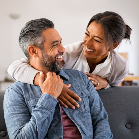 Man sitting on couch smiling at his long-term partner as she drapes her arm around him and looks at him lovingly