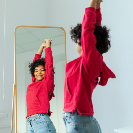 Woman stands with her arms raised smiling as she admires herself in a mirror